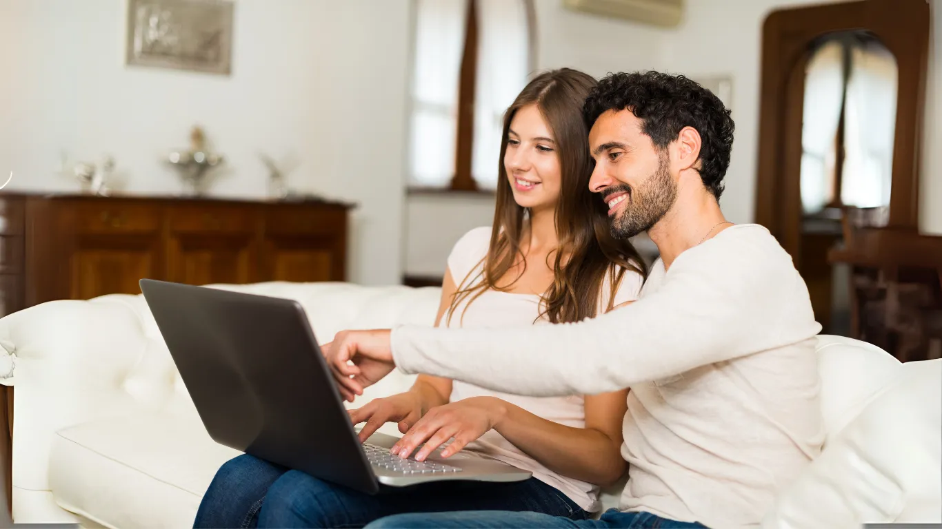 Uma mesa redonda com cadeiras em volta dela, na direita tem uma janela sem cortinas e na esquerda tem duas estantes com algumas decorações e livros.
